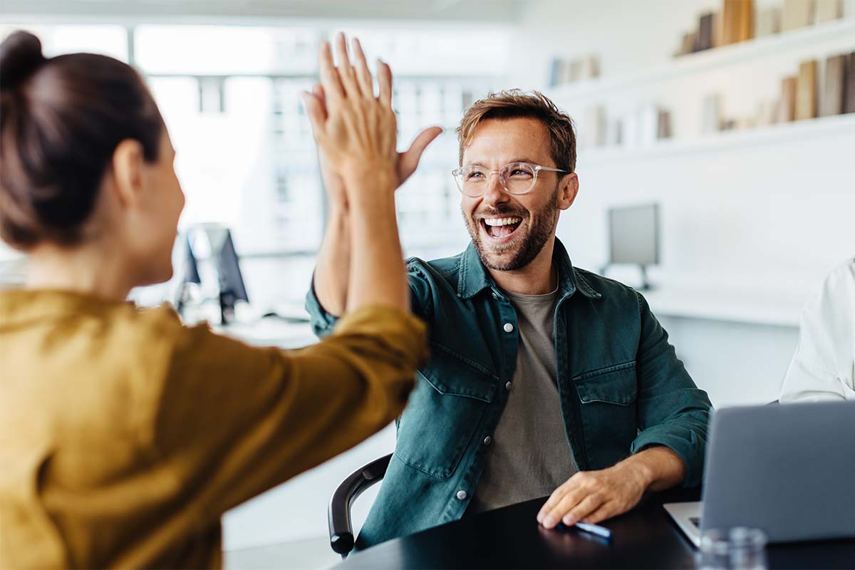 Zwei Kollegen geben sich im Sitzen ein High-Five am Büroarbeitsplatz.